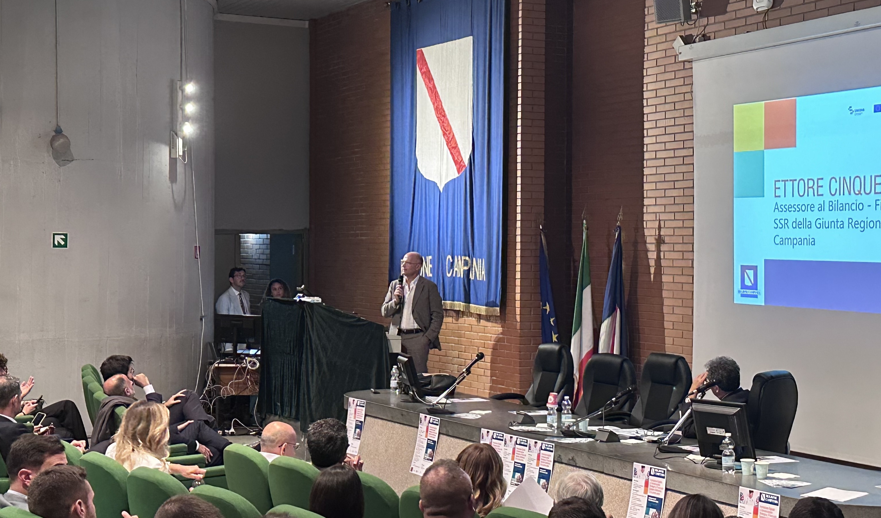 Uomo in piedi, con un microfono, mentre interviene di fronte a un pubblico 